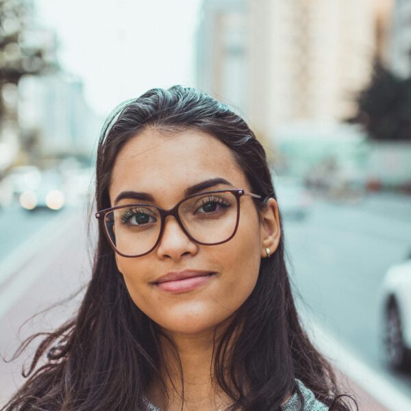 A confident woman wearing glasses smiles subtly while standing on an urban street with blurred background.